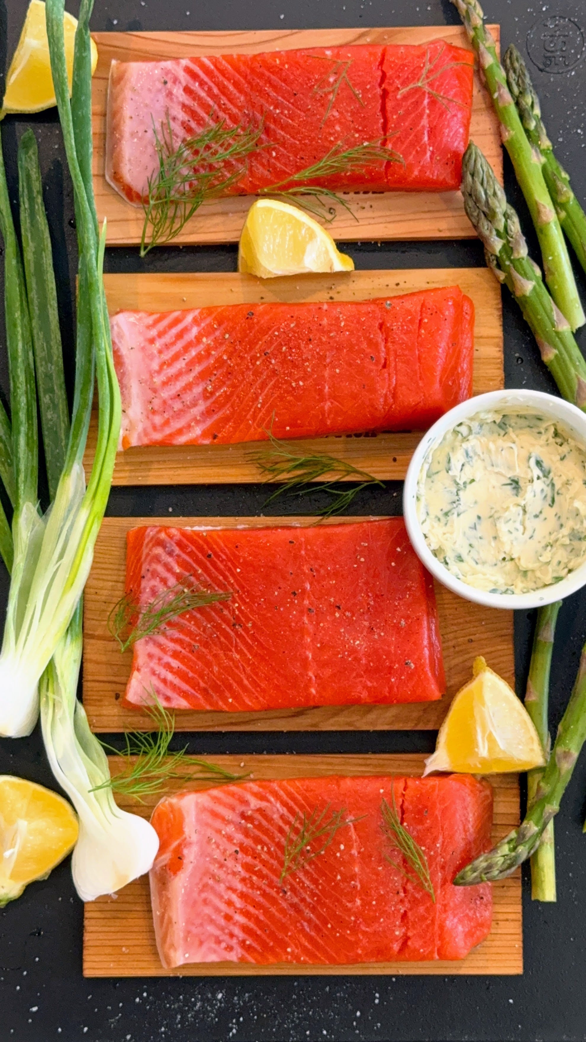 Raw salmon fillets on cedar planks with lemon, asparagus, and a small bowl of compound butter.