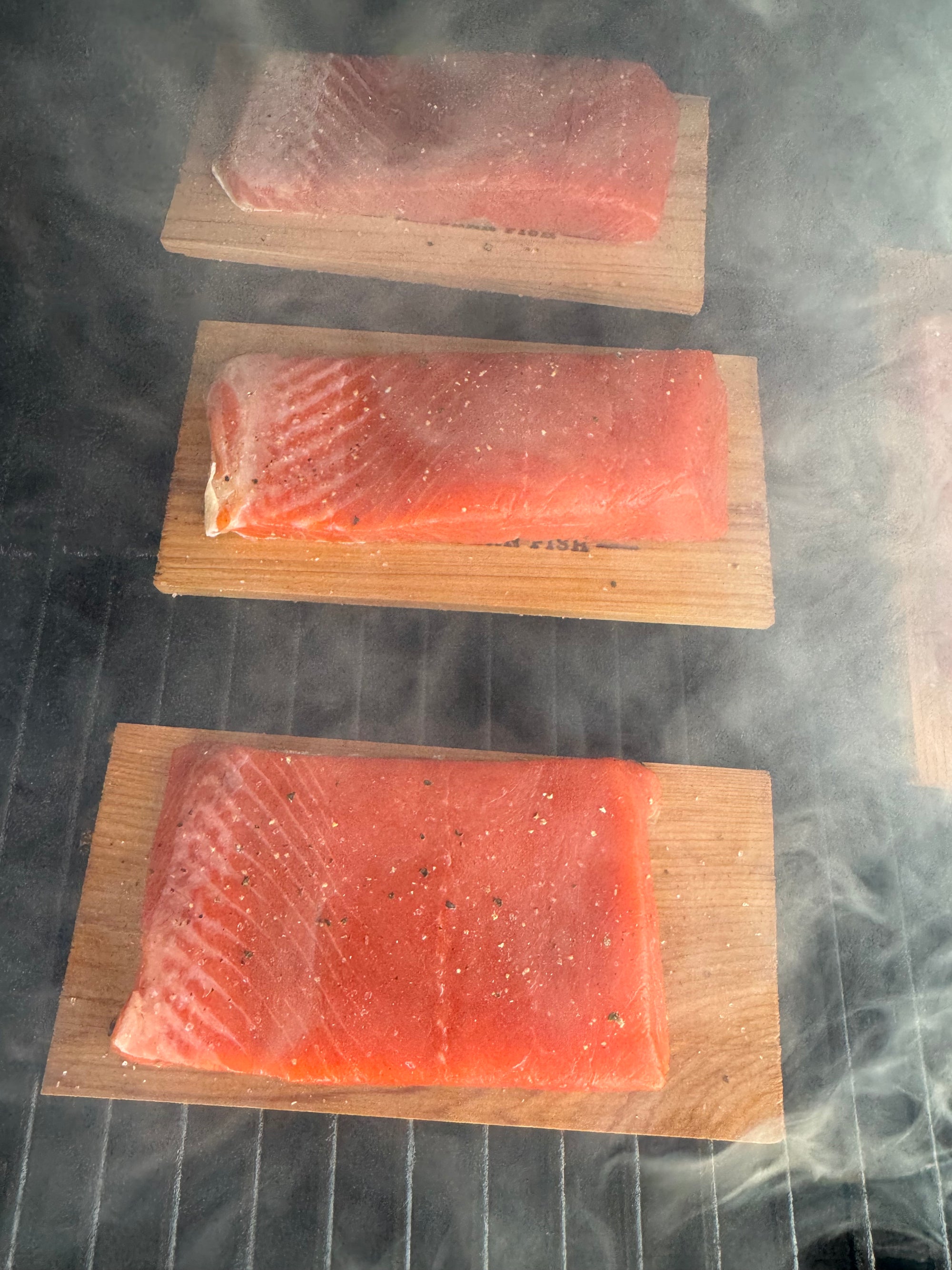 Three raw salmon fillets on cedar planks with smoke in the background