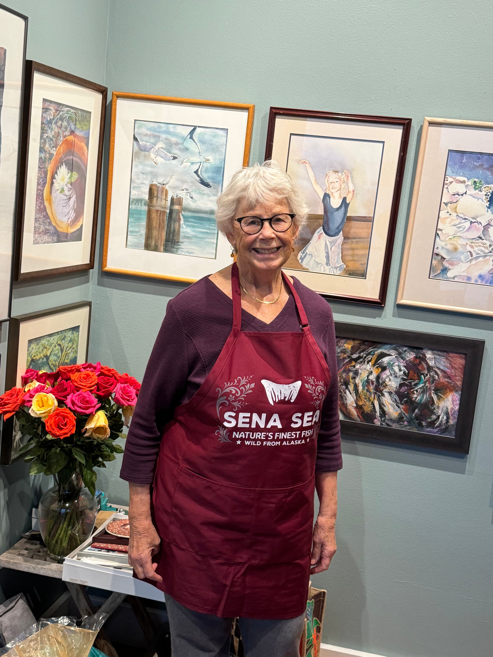 Woman wearing a maroon apron with 'Sena Sea' branding in an art gallery setting.