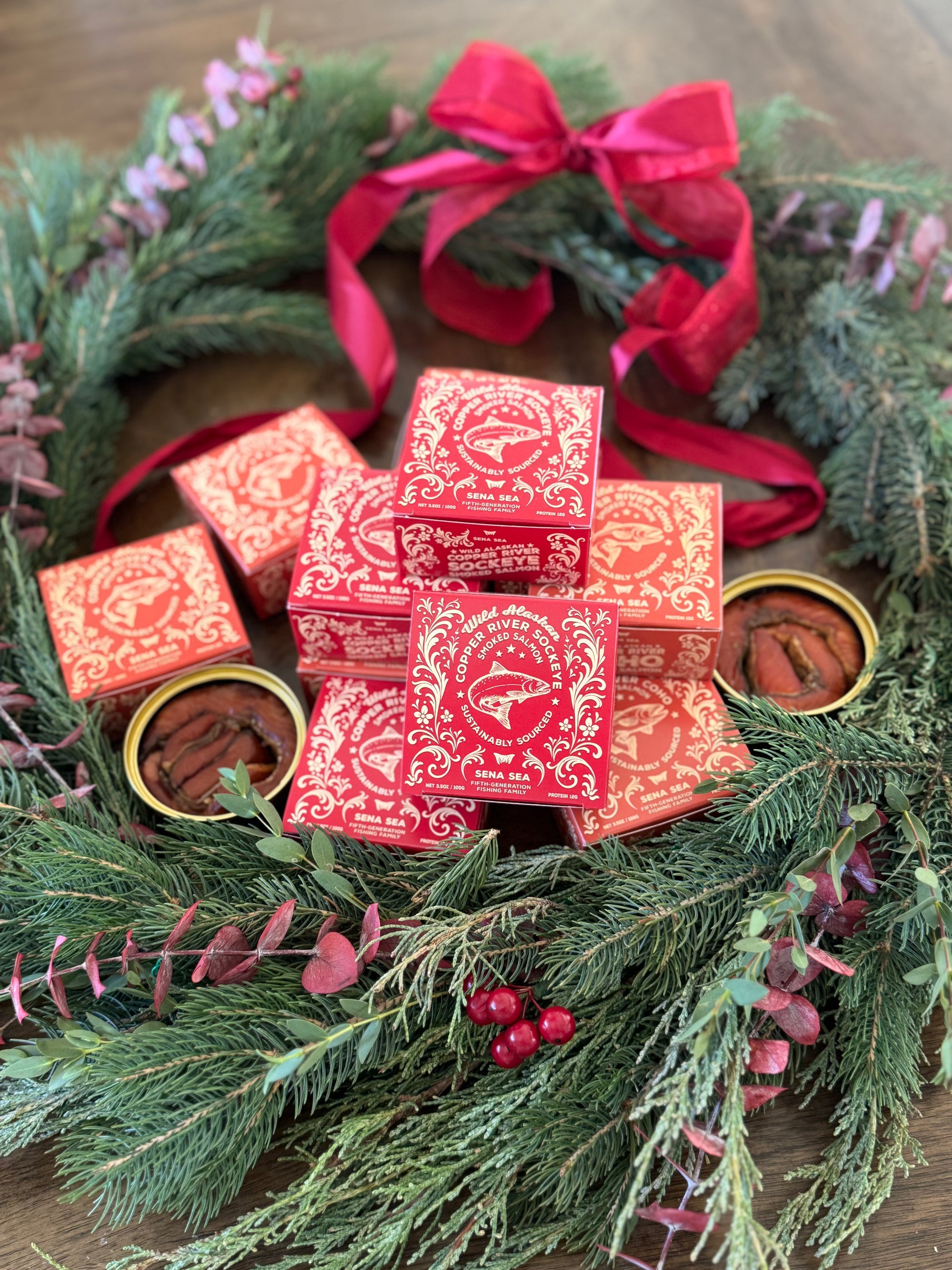 Red boxes of smoked Copper River salmon with gold accents on a festive green wreath with red ribbons and pine needles.