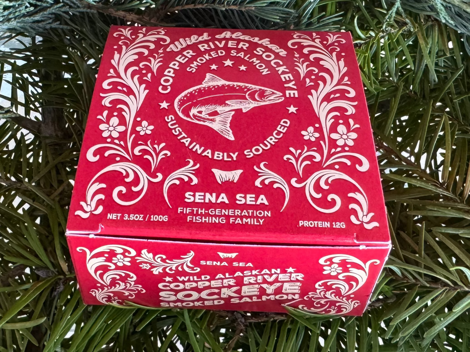 Red box of smoked copper river sockeye salmon with white text and rosmaling design on a green leafy background