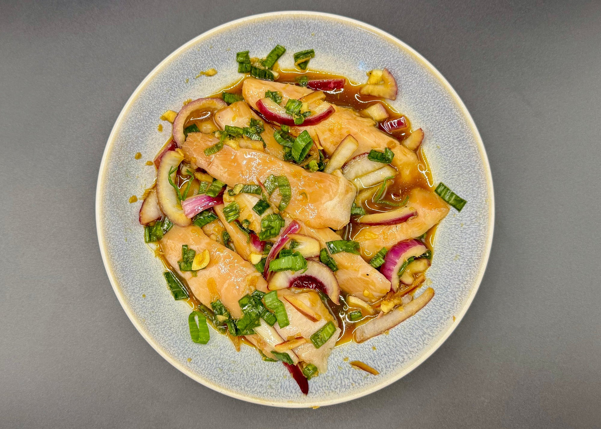 A top-down view of marinated sablefish tips on a light blue-gray speckled ceramic plate, set against a solid dark gray background. The sablefish (black cod) tips are mixed with sliced red onion, green onions.