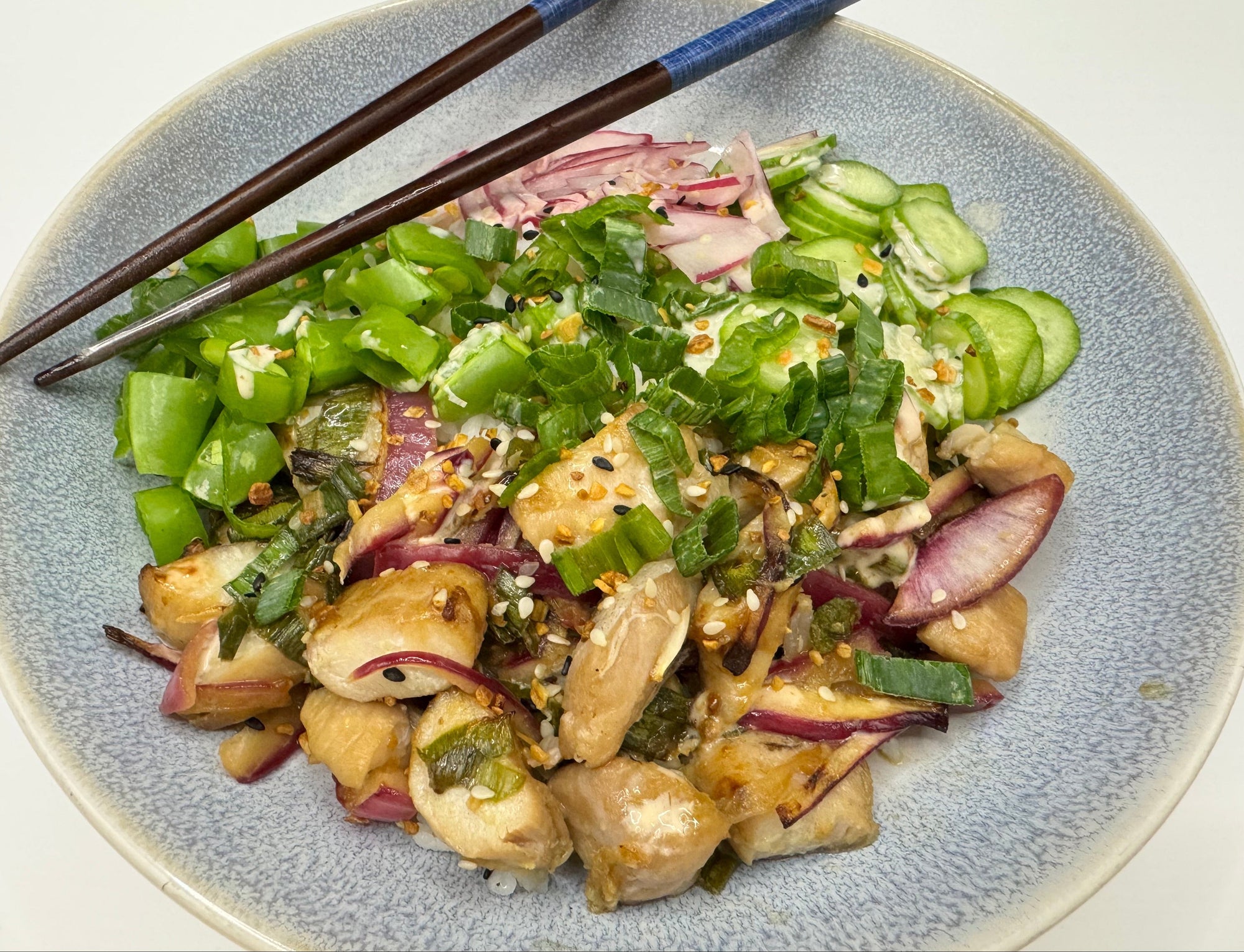 A close-up of a prepared poke bowl on a speckled light blue-gray plate, with chopsticks resting across the top. The poke bowl contains sliced cucumbers, chopped green onions, red onion, and marinated Sablefish (black cod) pieces.